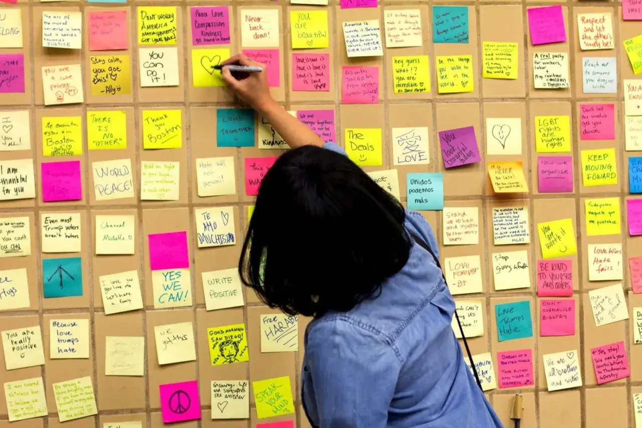 Una persona escribe en un mural de notas adhesivas de colores, un workshop que es un espacio de expresión y unidad.