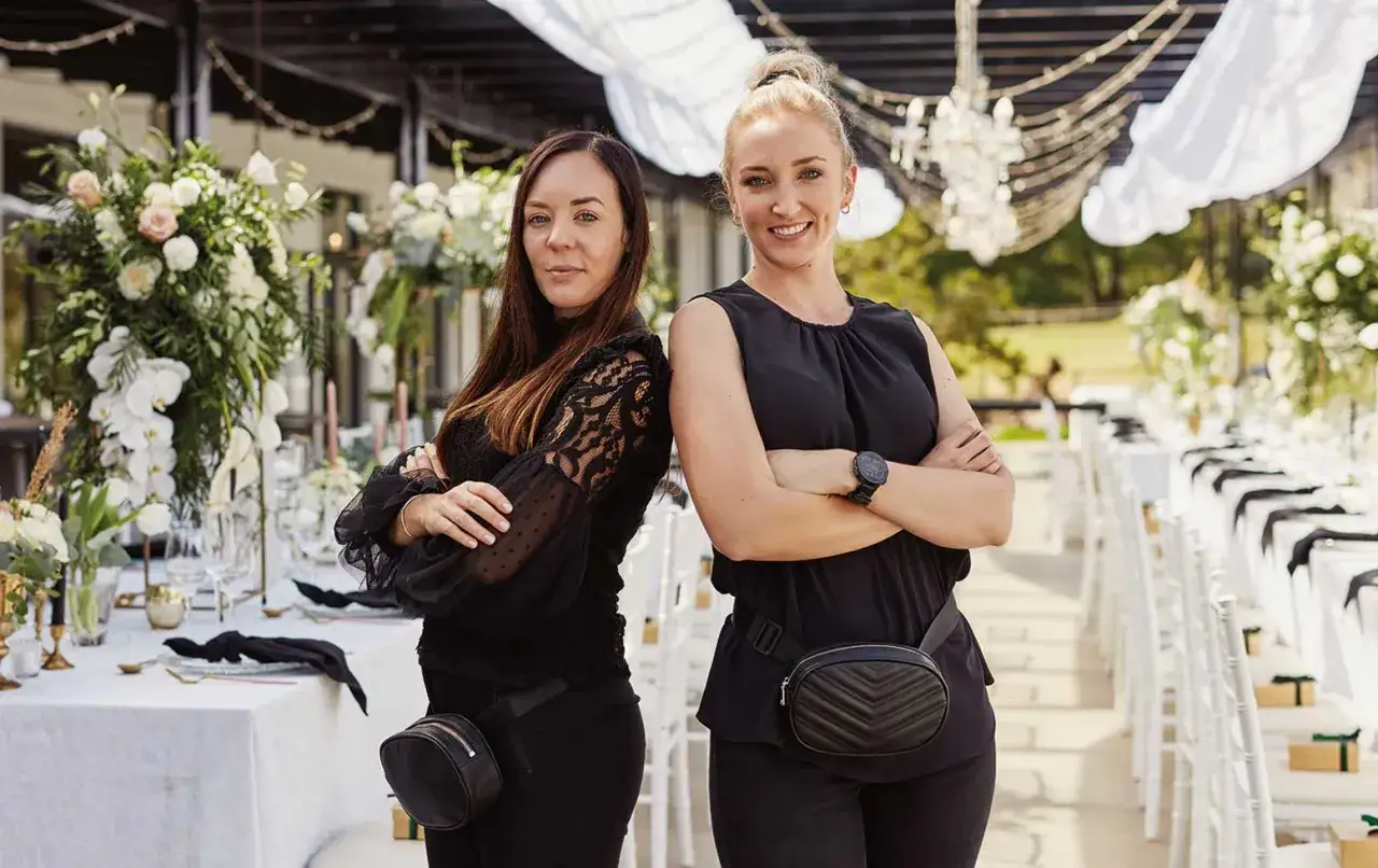 Dos organizadoras de eventos, vestidas de negro, supervisan la preparaci&oacute;n de una boda al aire libre, mostrando las distintas fases de un evento.