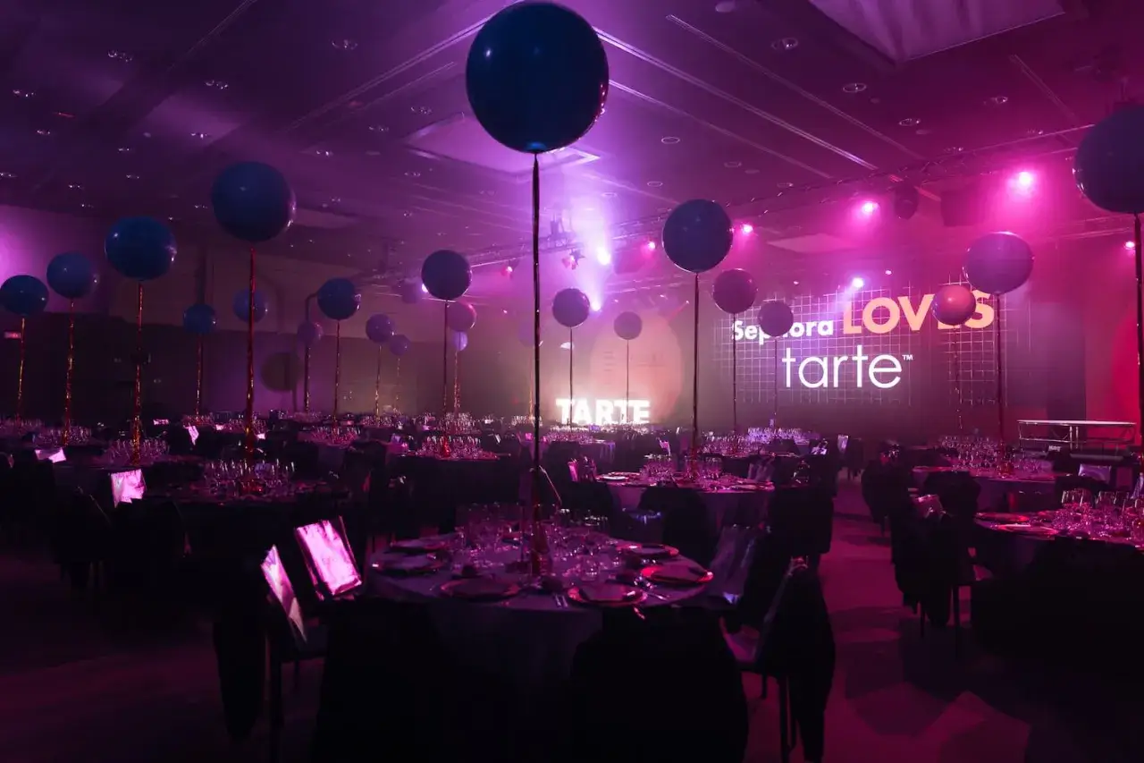 Sal&oacute;n de eventos de marca con mesas puestas, globos flotando y pantalla gigante que anuncia 