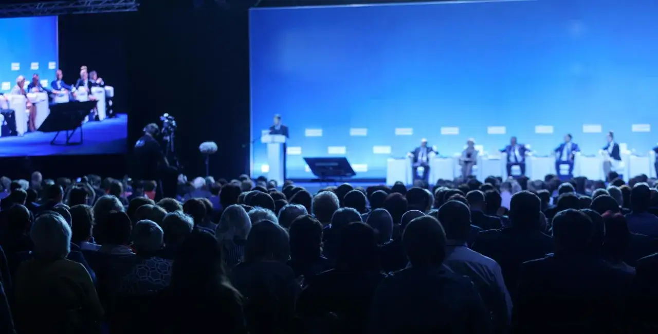 Un público numeroso asiste a un congreso. En el escenario, ponentes discuten temas importantes ante una gran pantalla azul.