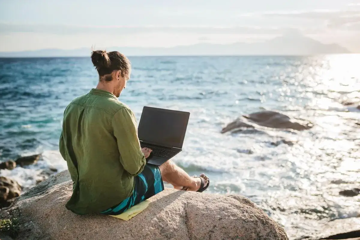 Hombre con portátil en roca junto al mar, disfrutando de trabajos para nómadas digitales.