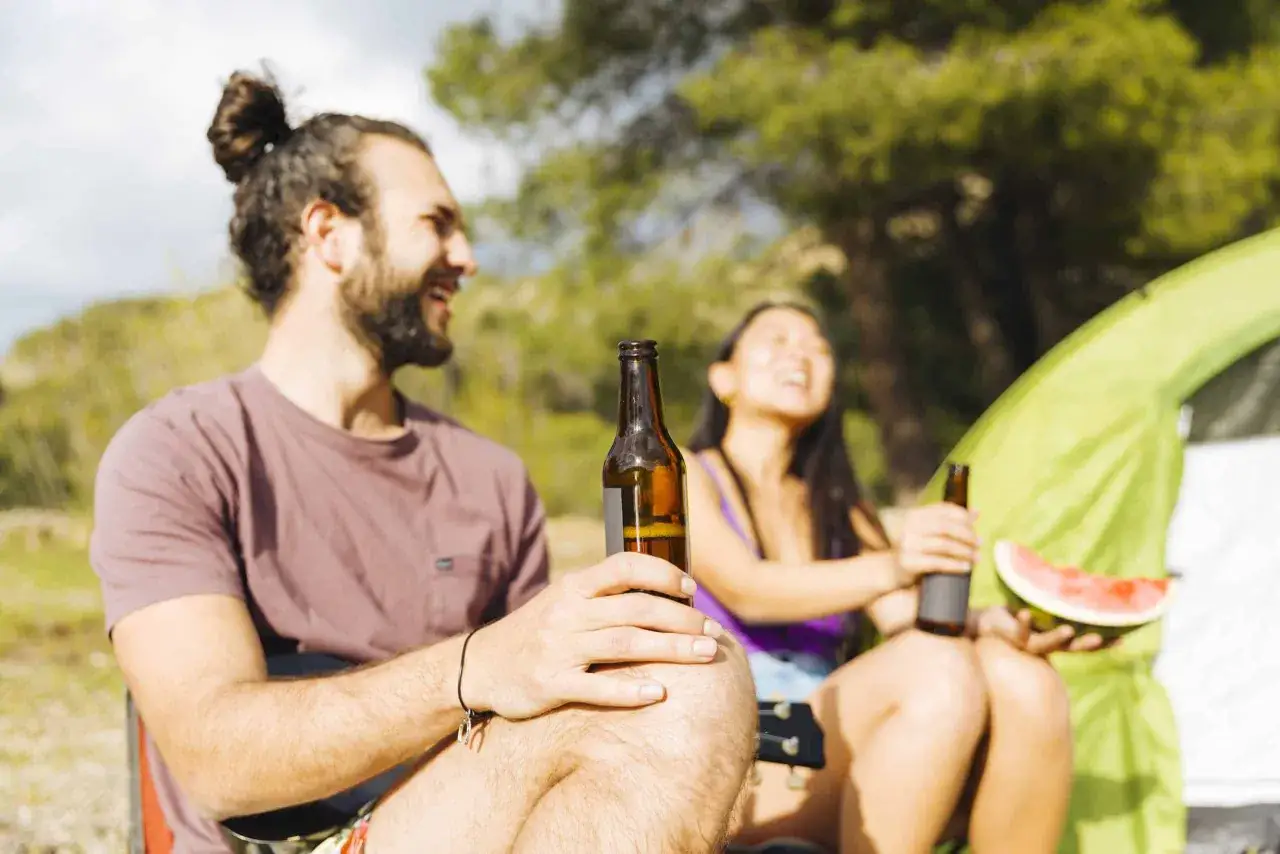 Pareja riendo con cervezas y sand&iacute;a, disfrutando de un d&iacute;a al aire libre, un ejemplo de eventos relajados.